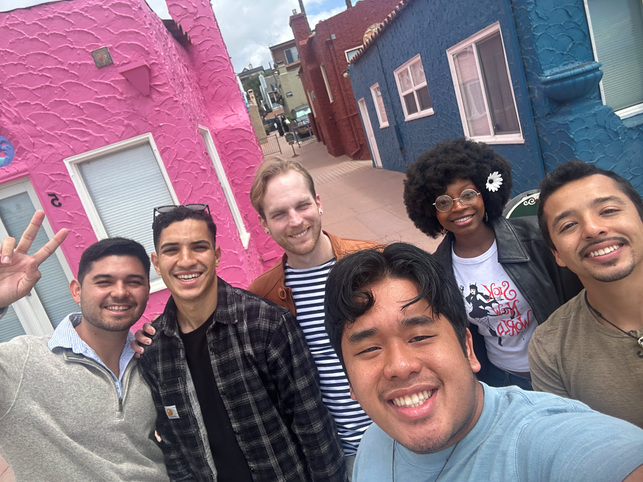 Six smiling students of a range of ethnicities pose for a candid shot in front of two brightly colored homes
