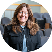 Woman wearing dark blue jacket, light blue shirt, and long hair, smiling while standing in a classroom.