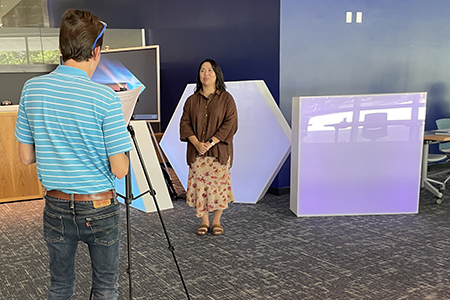 one person points tripod and camera in front of another who is standing in front of a bright large shape installation