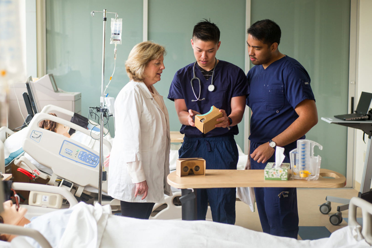 health students in scrubs look at clipboard with professor in lab coat. Clinic setting with dummy in hospital bed in foreground.